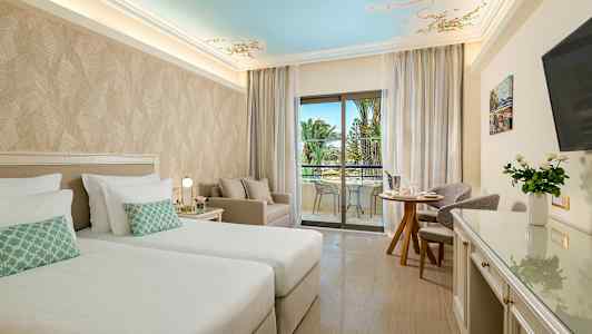 Hotellihuone, jossa kaksi vuodetta, istuinryhmä ja parveke näkymällä palmuihin. (Tekoälyn luoma teksti)