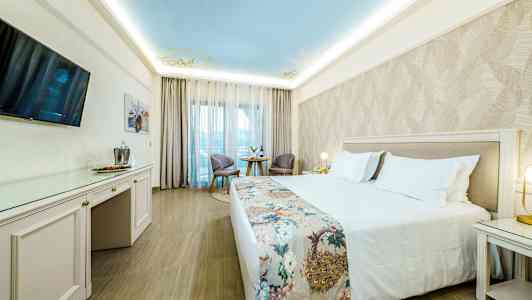 Hotellihuone, jossa on leveä parivuode ja koristeltu peitto, kaksi tuolia ja pöytä suuren ikkunan ääressä. (Tekoälyn luoma teksti)