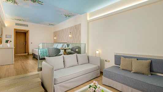 Valoisa hotellihuone, jossa istuinalue, sänky ja koristeltu katto. (Tekoälyn luoma teksti)