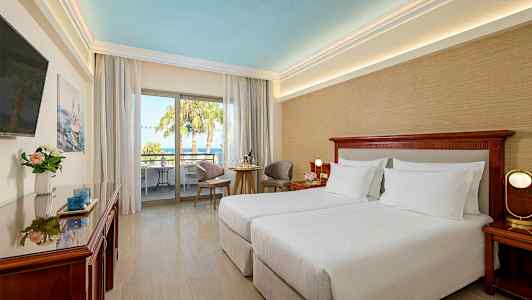 Hotellihuone kahdella erillisvuoteella, istuinryhmä ja parveke, josta on näkymä palmuihin. (Tekoälyn luoma teksti)