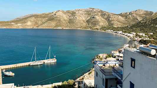 Näkymä rannikolle, jossa pieniä rakennuksia ja vuoria taustalla. Veneet ovat kiinnitettyinä laituriin kirkkaassa vedessä. (Tekoälyn luoma teksti)