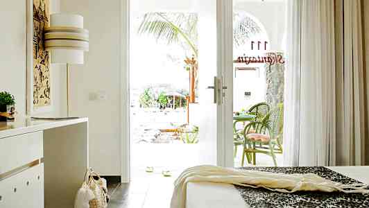 Hotellihuone, jonka avoimesta lasiovesta näkyy terassi, jossa on vihreitä tuoleja. (Tekoälyn luoma teksti)