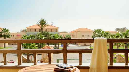 Näkymä parvekkeelta, jossa on pöytä ja pyyhe parvekkeen kaiteen päällä, taustalla näkyy rakennuksia ja palmuja. (Tekoälyn luoma teksti)