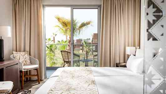 Hotellihuone, jossa on suuri sänky, istuma-alue ja parveke näkymällä palmupuihin. (Tekoälyn luoma teksti)