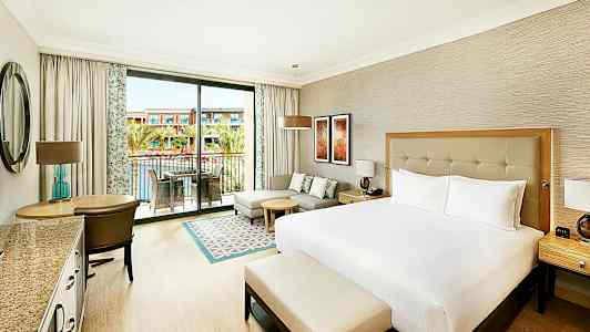 Hotellihuone, jossa on iso sänky, oleskelutila ja parveke näköalalla uima-altaalle ja rakennuksiin. (Tekoälyn luoma teksti)