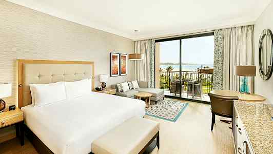 Hotellihuone, jossa on suuri sänky, oleskelutila, työpöytä ja patio, josta on näkymä merelle. (Tekoälyn luoma teksti)