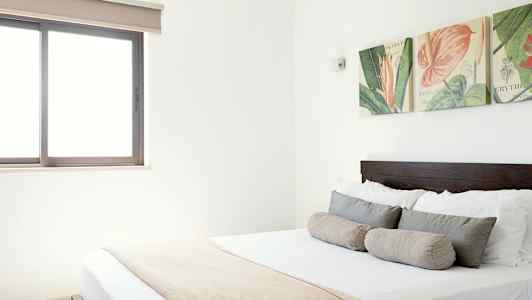 Kuva vaaleasta makuuhuoneesta, jossa on parisänky, koristeelliset tyynyt ja seinäkoristeet sängyn yläpuolella. (Tekoälyn luoma teksti)