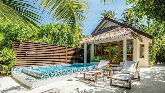 Bungalow, jossa olkikatto, pieni uima-allas ja aurinkotuoleja, ympäröitynä vehreydellä. (Tekoälyn luoma teksti)