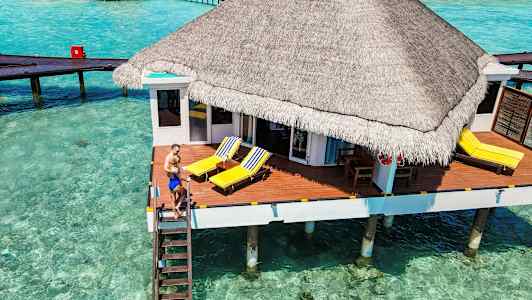 Bungalow paalujen päällä kirkkaan turkoosin veden yläpuolella, jossa aurinkoterassi ja rantatuolit. (Tekoälyn luoma teksti)