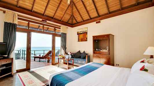 Hotellihuone, jossa on suuri sänky, puinen pöytä ja sohva, näkymä merelle lasiovien kautta. (Tekoälyn luoma teksti)