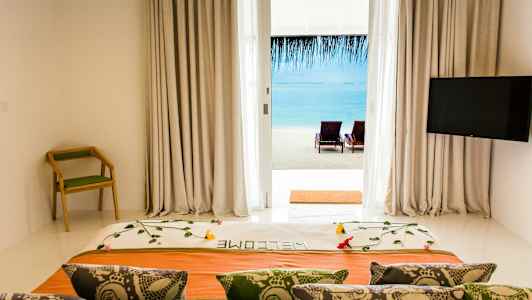 Näkymä hotellihuoneesta, jossa on koristeltu sänky, selkänojallista tuolia ja televisio, rannalle ja merelle lasioven kautta. (Tekoälyn luoma teksti)