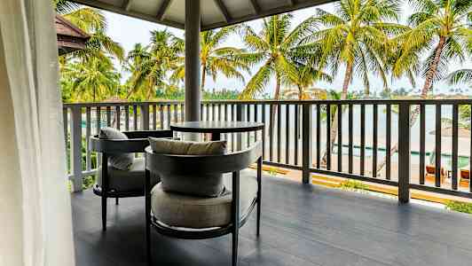 Kuva ulkoterassista, jossa on kaksi harmaata istuinta ja pöytä. Terassilta on näkymä palmupuiden yli rannalle. (Tekoälyn luoma teksti)