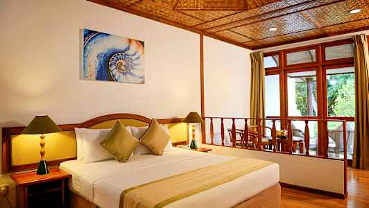 Hotellihuone, jossa iso sänky, kaksi yöpöytää lampuilla ja istuinryhmä ikkunan vieressä. (Tekoälyn luoma teksti)