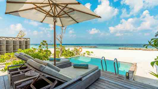 Kuva aurinkoterassista aurinkovarjolla ja aurinkotuoleilla, altaan vieressä, taustalla valkoinen hiekkaranta ja meri. (Tekoälyn luoma teksti)