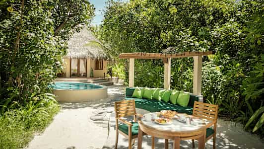 Puutarha, jossa vehreitä kasveja, pyöreä uima-allasalue, olkikattoinen talo ja istuinryhmä pöytineen ja tuoleineen. (Tekoälyn luoma teksti)