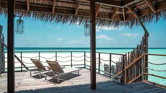 Kaksi aurinkotuolia puisella laiturilla, näkymä täynnä kirkasta sinistä merta olkikatoksen alla. (Tekoälyn luoma teksti)