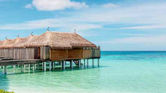Vesi-bungalowit, joissa olkikatot pylväillä turkoosin veden päällä ja kirkas taivas. (Tekoälyn luoma teksti)
