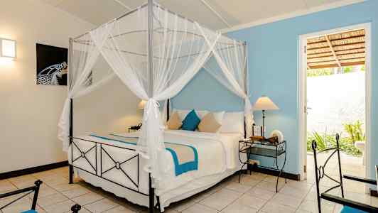 Makuuhuone, jossa on sälepohjainen sänky, valkoisia ja sinisiä tekstiilejä, kaksi yöpöytää lampuilla ja ovi, joka johtaa ulkoalueelle. (Tekoälyn luoma teksti)