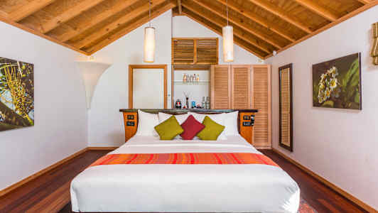 Makuuhuone, jossa on puurakenteinen katto, suuri vuode valkoisilla vuodevaatteilla ja värikkäitä koristeellisia tyynyjä, sekä sisustus sisältäen lamppuja ja seinäkoristeita. (Tekoälyn luoma teksti)