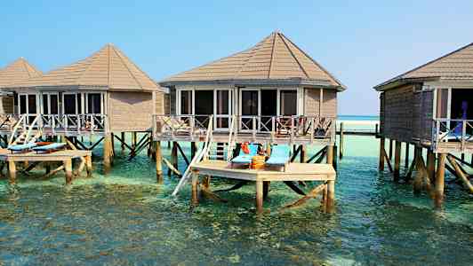 Kuvassa on paalujen päällä olevia mökkejä kirkkaan veden yllä. Terassilla on aurinkotuoleja. (Tekoälyn luoma teksti)