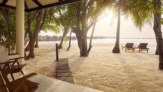 Näkymä terassilta hiekkarannalle, jossa on aurinkotuoleja, puita ja veden päällä olevia bungaloweja taustalla. (Tekoälyn luoma teksti)