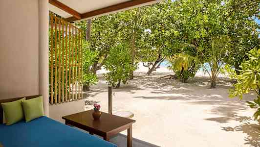 Näkymä terassilta, jossa on sininen sohva ja pöytä, jolla on maljakko, hiekan ja vehreän puuston ympäröimänä lähellä merta. (Tekoälyn luoma teksti)