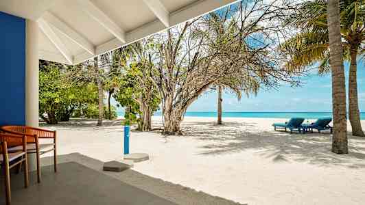 Hiekkaranta, jossa on muutamia aurinkotuoleja, palmuja ja sininen meri taustalla, nähtynä terassilta. (Tekoälyn luoma teksti)