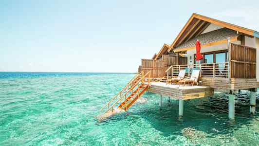 Valokuva bungalowista pylväiden päällä turkoosin veden yllä. (Tekoälyn luoma teksti)