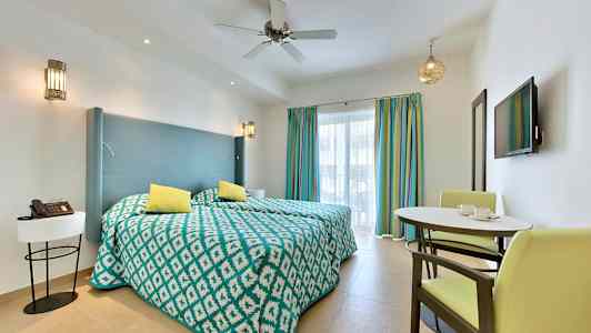 Hotellihuone, jossa on kaksi erillistä vuodetta, pieni ruokailupaikka ja seinätelkkari värikkäiden verhojen edessä. (Tekoälyn luoma teksti)