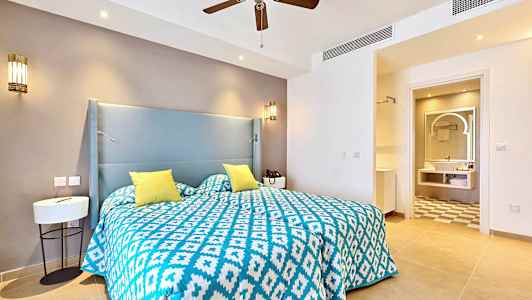 Hotellihuone, jossa on sinivalkoinen kuvioitu peitto ja keltaiset tyynyt sängyllä. Taustalla näkyy kylpyhuone. (Tekoälyn luoma teksti)