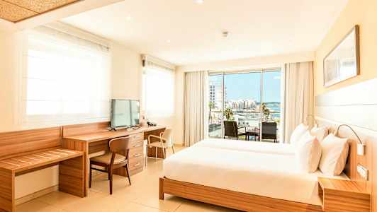 Valoisa hotellihuone, jossa on suuri sänky, työpöytä ja televisio sekä parveke, jolta on näkymä kaupungille ja merelle. (Tekoälyn luoma teksti)