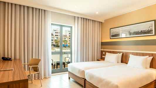 Hotellihuone, jossa on kaksi erillistä vuodetta, kirjoituspöytä ja lasiovellinen parveke. (Tekoälyn luoma teksti)