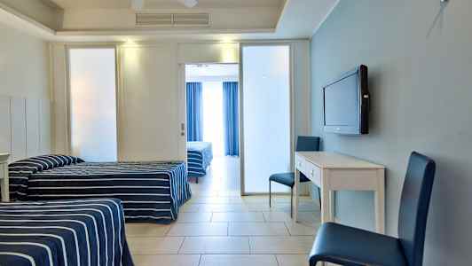 Kaksi yhden hengen vuodetta, joissa on sinivalkoiset raidalliset päiväpeitteet, valkoinen kirjoituspöytä, kaksi sinistä tuolia ja seinään kiinnitetty TV, sekä lasiovi takana olevaan huoneeseen. (Tekoälyn luoma teksti)