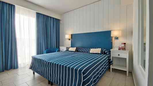 Hotellihuone, jossa on parisänky sinivalkoraidallisilla vuodevaatteilla, sininen sängynpääty ja valkoiset huonekalut. (Tekoälyn luoma teksti)