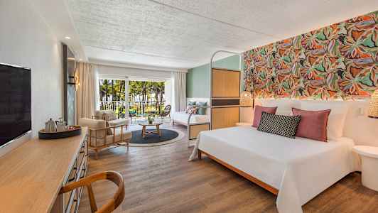 Hotellihuone, jossa on parivuode, oleskelutila, työpöytä ja parveke näkymällä palmuihin ja puutarhaan. (Tekoälyn luoma teksti)