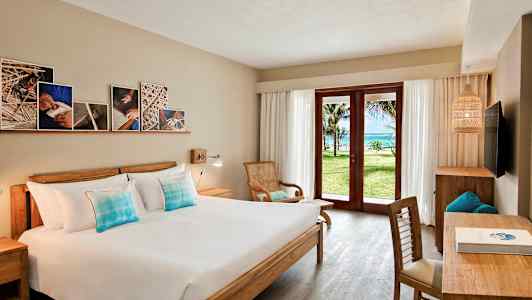 Hotellihuone, jossa on parivuode, puisia huonekaluja ja lasiovet, jotka avautuvat vihreälle puutarhalle ja merinäköalalle. (Tekoälyn luoma teksti)