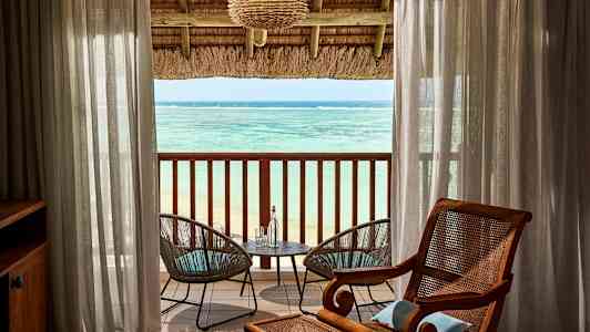 Näkymä merelle huoneesta, jossa on katettu parveke. Läpinäkyvien verhojen välissä on kaksi puutarhatuolia pöydän ääressä. (Tekoälyn luoma teksti)