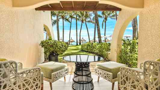 Näkymä terassilta, jolla on tuoleja ja pöytä, puutarhaan, jossa on palmuja ja ranta taustalla. (Tekoälyn luoma teksti)