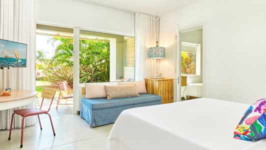 Vaalea hotellihuone, jossa on sänky, oleskelutila ja pöytä. Näkymä puutarhaan näkyy lasiovien läpi. (Tekoälyn luoma teksti)