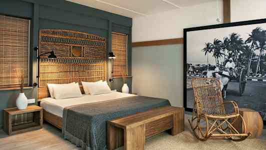 Hotellihuone, jossa on parivuode, puisia yksityiskohtia ja rottinkituoli. Suuri mustavalkoinen kuva koristaa seinää. (Tekoälyn luoma teksti)