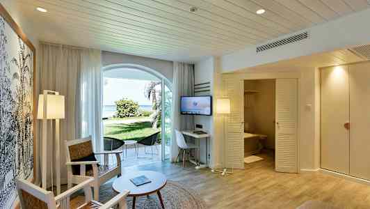 Hotellihuone, jossa istuimet, pöytä ja TV. Ovista pääsee kylpyhuoneeseen, ja ikkunoista avautuu näkymä vihreälle puutarhalle ja rannalle. (Tekoälyn luoma teksti)