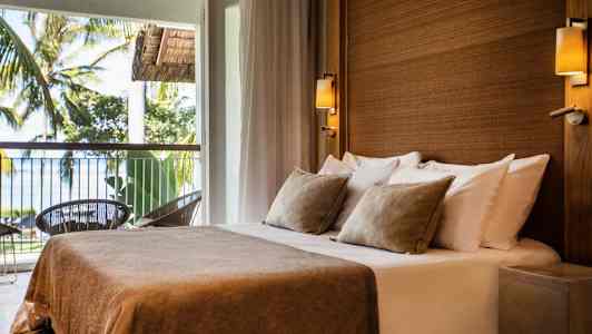 Huone, jossa on parivuode beigeillä vuodevaatteilla ja tyynyillä, sijaitsee parvekkeella, jolta on näkymä puihin ja mereen. (Tekoälyn luoma teksti)