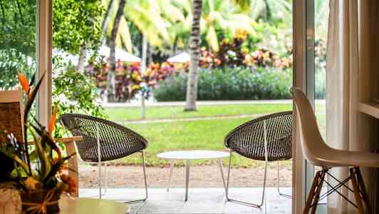 Näkymä huoneesta avoimen oven kautta patiolle, jossa on kaksi tuolia ja pöytä, ympäröitynä viheralueilla ja palmupuilla. (Tekoälyn luoma teksti)