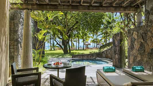 Terassi, jossa on istuimia ja aurinkotuoleja pienen altaan vieressä, ympäröitynä kasveilla ja näkymällä rannalle ja palmuihin. (Tekoälyn luoma teksti)