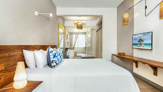 Kuva modernista hotellihuoneesta, jossa on puukalusteet, sänky valkoisilla lakanoilla ja värikkäillä tyynyillä sekä suuri ikkuna taustalla. (Tekoälyn luoma teksti)