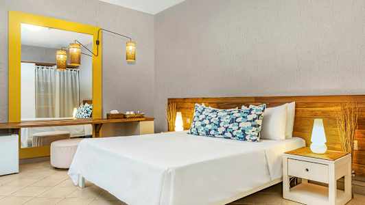 Hotellihuone, jossa on sänky koristetyynyjen kanssa, yöpöytä lampulla ja suuri keltareunainen peili. (Tekoälyn luoma teksti)