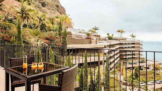 Kuva parvekkeesta, jossa on rottinkikalusteet ja pöytä, jolla on kaksi lasia ja karahvi. Näkymä istutusalueeseen, rakennuksiin ja merelle. (Tekoälyn luoma teksti)