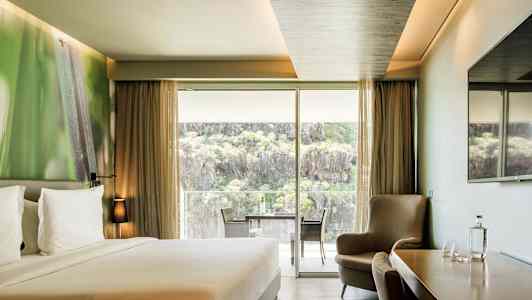 Hotellihuone, jossa on parivuode, istuinryhmä, pöytä tuoleineen sekä suuri lasiovi, joka avautuu parvekkeelle ja tarjoaa näköalaa luontoon ja kallioihin. (Tekoälyn luoma teksti)