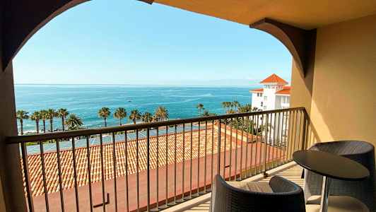 Näkymä parvekkeelta, jossa on kaksi tuolia ja pöytä, kohti merta ja palmuja. (Tekoälyn luoma teksti)