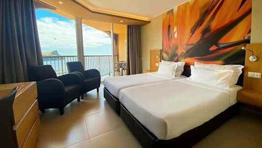 Hotellihuone, jossa kaksi valkoista sänkyä, värikäs seinämaalaus ja parveke merinäköalalla. (Tekoälyn luoma teksti)